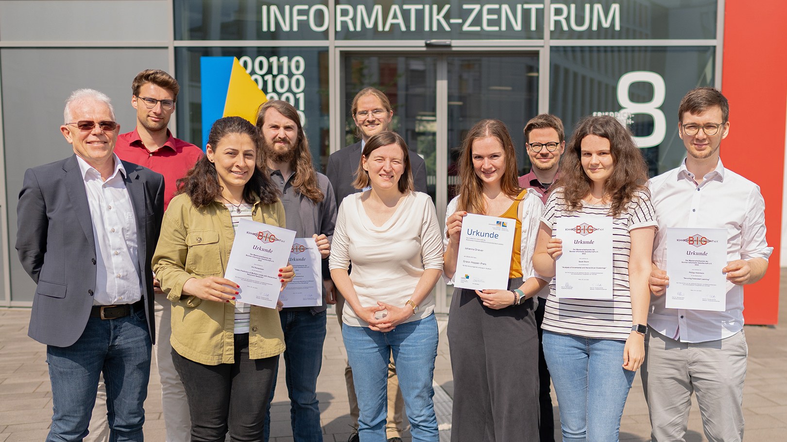 Eine Wissenschaftlerin und ein Wissenschaftler arbeiten hinter einer Glasfassade und mischen Chemikalien mit Großgeräten.